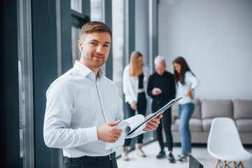 Fototapeta premium Man in formal clothes standing with documents in front of young successful team that working and communicating together indoors in office