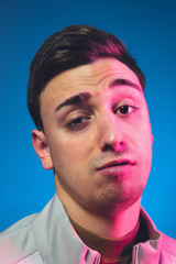 Fototapeta premium Gremaces, looking on. Close up caucasian man's portrait isolated on blue studio background in pink neon light. Beautiful male model. Concept of human emotions, facial expression, sales, ad. Copyspace.