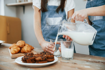 Close up view of two little girls in blue chef uniform that pouring milk into glasses on the kitchen with cookies on table
