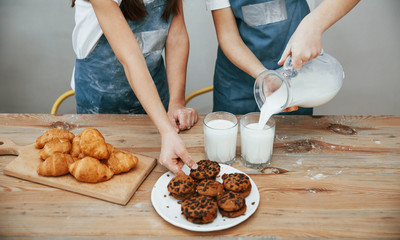 Close up view of two little girls in blue chef uniform that pouring milk into glasses on the kitchen with cookies on table