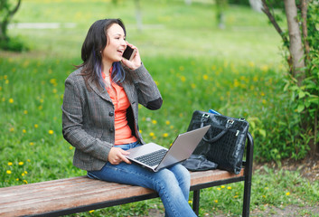 Young freelancer girl sits on the bench in park, working on laptop and talking on phone