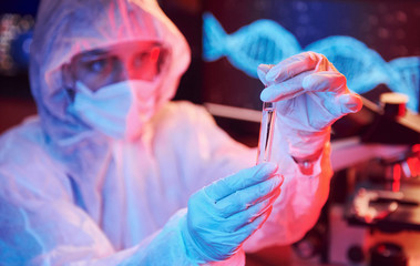 Nurse in mask and white uniform, holding tube with liquid and sitting in neon lighted laboratory...