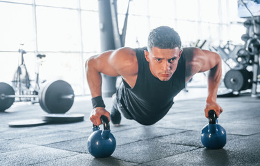 Strong young man in sportive clothes doing push ups in gym by using dumbbells