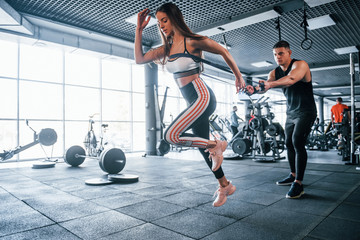 Young sportive people doing exercises together in the gym. Woman running when man holding her by elastic tape