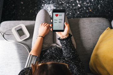 Young woman measures blood pressure sitting on sofa at home with smartphone connected to device -...