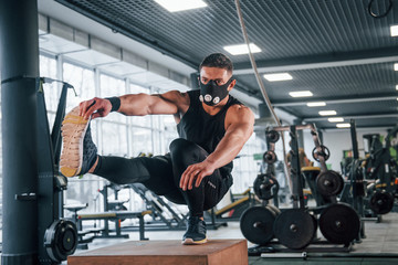 Fototapeta na wymiar Strong young man in sportive clothes and scary mask doing push ups on one leg in gym