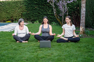 Mother, grandmother and teen daughter practicing online yoga class outdoors in garden at quarantine isolation period during coronavirus pandemic. Family doing sport together online from home. 