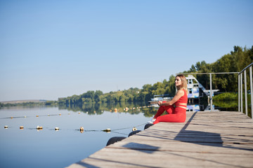 Young blond woman, wearing red fitness outfit sitting on wooden pier dock in summer morning. Healthy active life concept. Portrait of girl resting after training exercise outside in the city.