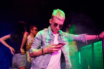 Guy standing with smartphone in front of oung people that having fun in night club with colorful...