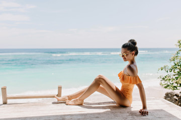 Refined slim young woman posing on the ground with sky on background. Pleasant tanned girl in sunglasses and stylish swimsuit chilling on ocean coast with shy smile.