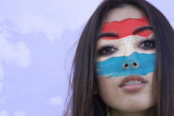 Flag of Luxemburgo painted on a face of a smiling luxembourgish young woman. Copyspace.