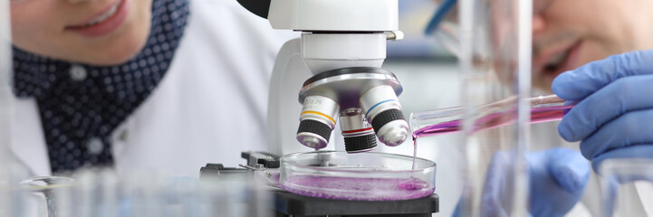 Close-up view of biochemist adding bright violet substance in glass container. Female chemist in...