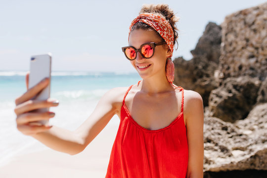 Stunning young lady in red attire using phone for selfie at wild beach. Adorable white girl in sparkle sunglasses taking picture of herself while resting at ocean.