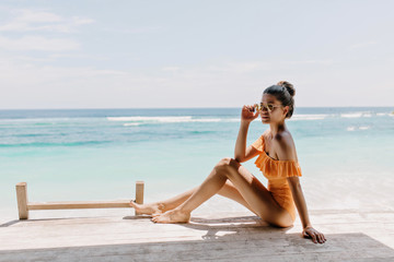 Barefooted amazing woman sitting on the ground on sea background. Magnificent tanned lady in swimsuit touching her sunglasses while posing at exotic resort and enjoiyng wave sounds.
