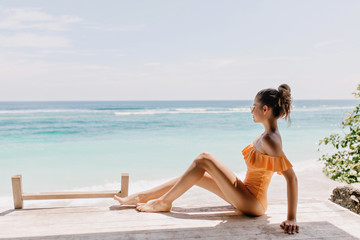 Pleasant woman in romantic swimsuit sitting on the ground and looking at horizon. Outdoor photo of slim white female model chilling at sea coast under clear sky.