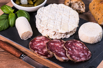 Typical european aperitif platter with goat cheese, dry smoked sausage, pickles and bread on wooden background. France 2020.