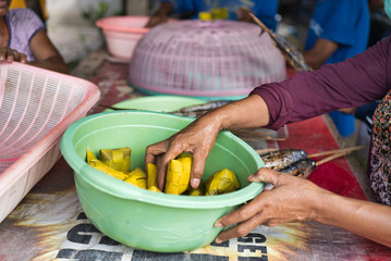Woman's hand holding a rice tamale wrapped in corn husks from a plastic green bowl at a Southeast Asian market.