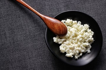 Closeup of kefir grains in black bowl on dark gray background with bamboo spoon. Macro, top view.