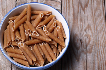 Primo piano di ciotola di ceramica contenente pennette biologiche integrali crude. Sfondo di legno grezzo con spazio per didascalia.