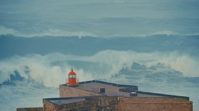Fantastic shot of a big wave coming towards the red lighthouse on a cliff in Nazare, Portugal. Popular tourist spot and travel destination for people who love extreme sports. Surfing on giant waves. 