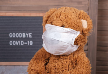 a cute Teddy bear in a medical mask sits at home against the background of a wind Board with the text in English goodbye covid-19. concept of the end of the coronavirus pandemic in the world