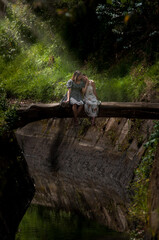 two young women with dresses sitting together on a rustic wooden bridge bathed in the sun on a river in the middle of nature