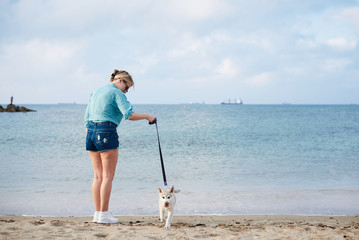 Young blond woman, wearing jeans shorts and turquoise blouse, standing at the beach by the sea, holding light grey white husky puppy with purple leash. Woman, walking with dog at seaside.
