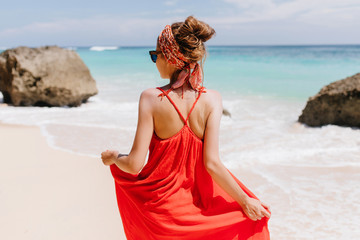 Photo from back of tanned gorgeous woman looking at sea. Outdoor portrait of amazing caucasian girl in red summer dress standing on sky background.