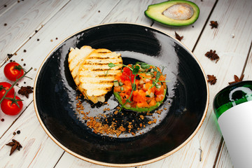 Toasted croutons and a salad of fresh tomatoes, herbs and pepper lie on a black plate. Near a wooden background are a bottle, avocado and cherry tomatoes.