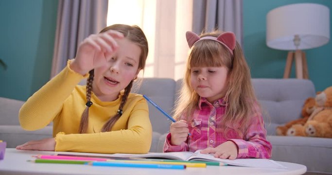 Joyful little Caucasian sisters staying at home alone and playing cheerfully together. Nice kids coloring picture with colorful pencils in cozy room. Happy children play and talking indoors.