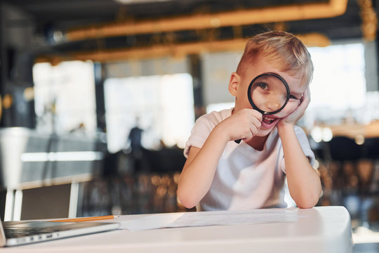 Smart child in casual clothes with laptop on table have fun with magnifying glass