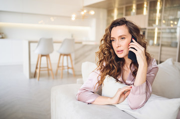 Beautiful young woman with curly hair in  pink pajamas at white couch in the morning. Lady speaking phone in scandinavian style living room interior. Distance work from home. Quarantine COVID-19