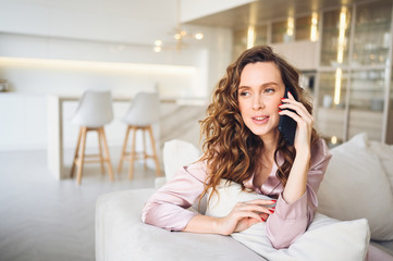 Beautiful young woman with curly hair in  pink pajamas at white couch in the morning. Lady speaking phone in scandinavian style living room interior. Distance work from home. Quarantine COVID-19