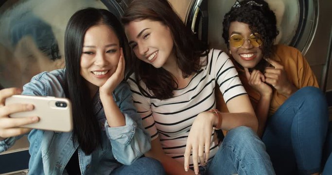 Three pretty mixed-races friendly girls at washing machines in washhouse smiling to smartphone camera, taking selfire photo. Multiethnic beautiful woman making pictures with phone in laundry service.