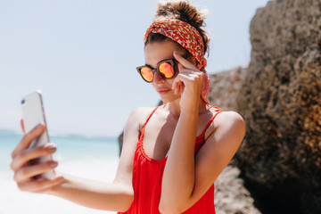 Serious european woman in sunglasses taking picture of herself on ocean background. Stunning tanned female model standing beside rocks and making selfie.