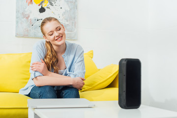 Selective focus of smiling woman near wireless speaker and laptop on coffee table in living room