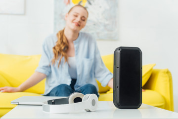 Selective focus of wireless speaker, headphones and laptop on coffee table near smiling woman on...