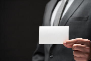 white blank business card closeup in businessman hand, gray suit, dark wall background