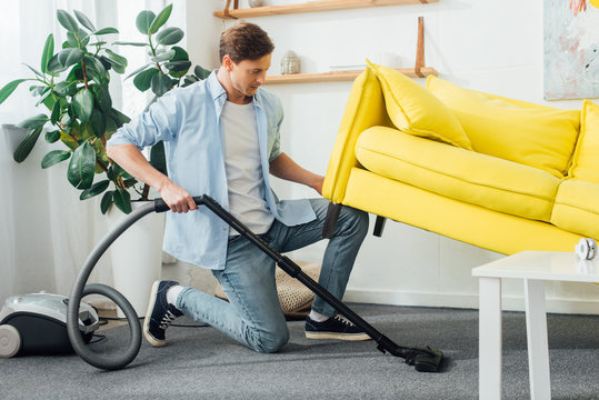 Side View Of Man Lifting Up Couch While Cleaning Carpet With Vacuum Cleaner