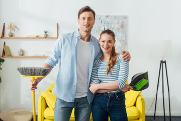 Handsome man smiling at camera while hugging girlfriend with scoop and broom in living room