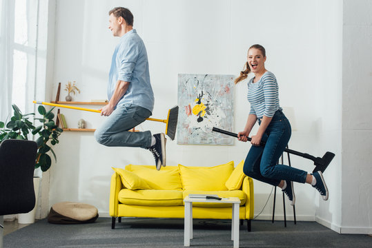 Side View Of Cheerful Couple Levitating On Brooms In Living Room