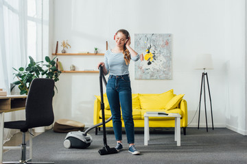Fototapeta na wymiar Attractive smiling woman in headphones cleaning carpet with vacuum cleaner at home
