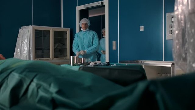 Two professional surgeons in uniforms, gloves, masks and hats enter the operating room. Surgical instruments and a patient with local anaesthesia are ready for a complex operation.