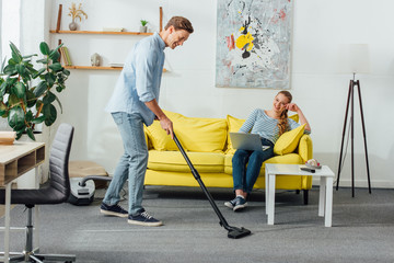 Smiling man cleaning carpet with vacuum cleaner near girlfriend using laptop on sofa at home