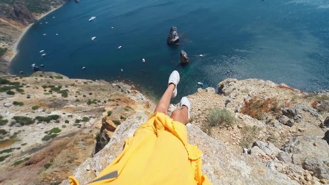 camera zooming out first person view of taned woman legs in yellow dress sitting on on cliff edge. amazing nature view of seascape with high limestone cliff over blue sea and clear deep blue sky