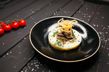 Meat on a plate of tender mashed potatoes on a black glossy plate. The black table is sprinkled with salt and decorated with cherry tomatoes.