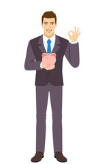 Smiling Businessman holding a piggy bank and showing a okay hand sign. Full length portrait of Businessman in a flat style.
