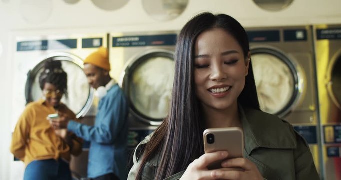 Close up of Asian pretty cheerful woman tapping and texting message on smarphone in laundry service room. Beautiful happy girl typing on phone and waiting for clothes to get clean in washhouse.