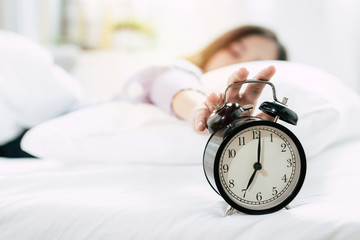 young asian woman sleeping on bed pressing snooze button on black vintage alarm clock at seven o'clock morning in bed room at home, lifestyle, good morning, healthy sleep and joyful weekend concept