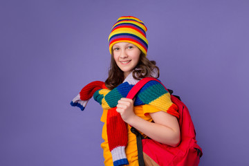 Fototapeta na wymiar Schoolgirl girl smiles in yellow T-shirt, knitted sweater and multi-colored hat with a backpack on her shoulder on a purple background